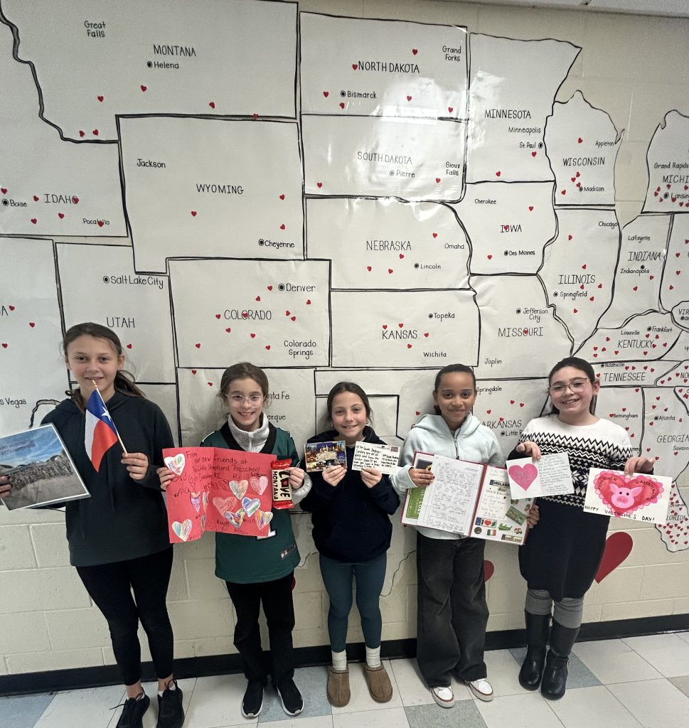 Five fifth grade students holding Valentine's Day cards standing in front of map of United States 