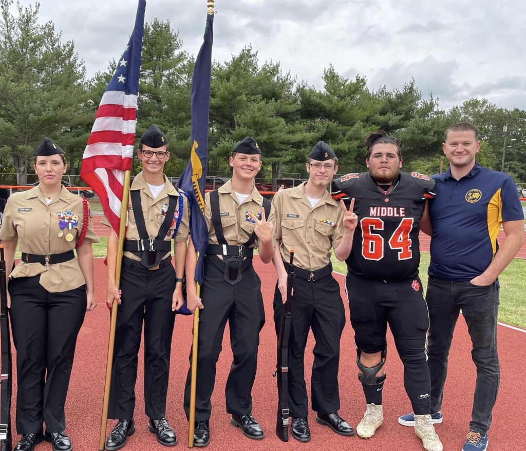 Four NNDCC cadets stand with senior football player and NNDCC instructor on sports field.